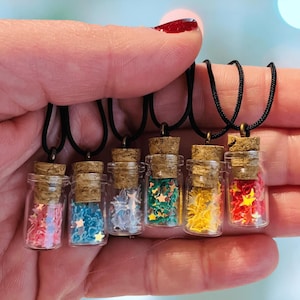 Puede incluir: Seis pequeños collares con botellas de vidrio y tapones de corcho, cada uno lleno de confeti en forma de estrella de colores. Las botellas están suspendidas de cordones negros. Los colores del confeti incluyen rosa, azul, blanco, verde, amarillo y rojo.
