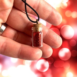 Puede incluir: Un vial de vidrio lleno de purpurina roja con un tapón de corcho, colgado de un collar de cordón negro.