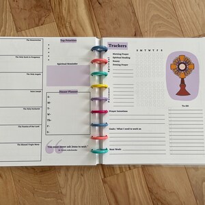 May include: A white planner with a purple and white design. The planner has a daily schedule, a section for top priorities, a dinner planner, and a section for trackers. The trackers include morning prayer, spiritual reading, rosary, evening prayer, prayer intentions, goals, and to-do list. The planner also has a quote that says "You must never ask Jesus to wait." The planner is closed and has a ring binder.