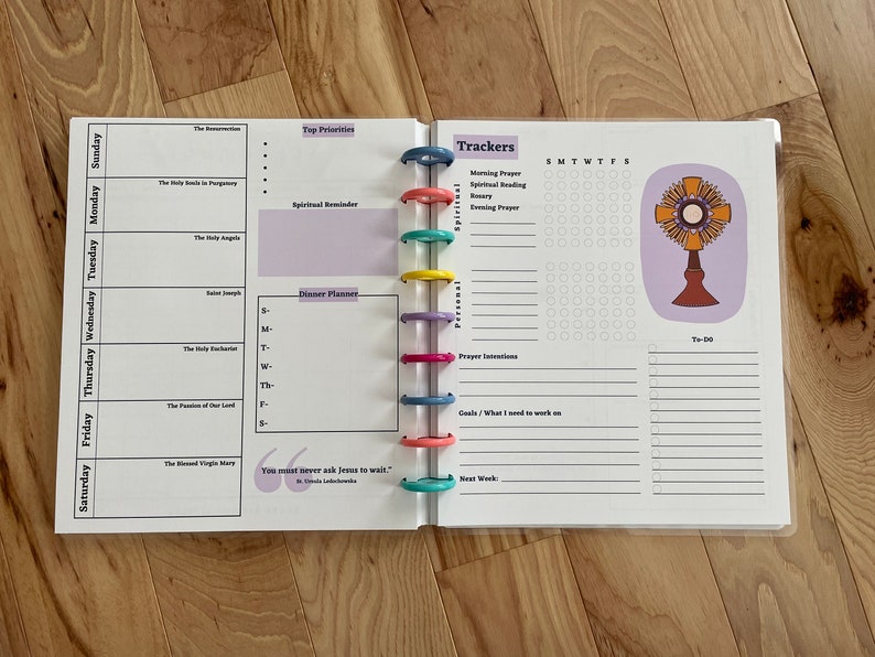 May include: A white planner with a purple and white design. The planner has a daily schedule, a section for top priorities, a dinner planner, and a section for trackers. The trackers include morning prayer, spiritual reading, rosary, evening prayer, prayer intentions, goals, and to-do list. The planner also has a quote that says "You must never ask Jesus to wait." The planner is closed and has a ring binder.