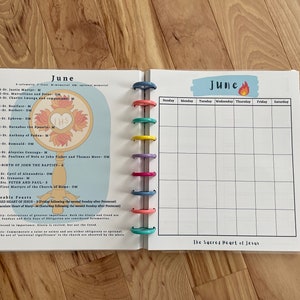 May include: A June calendar page with a colorful design. The left side features a religious image of a monstrance with the letters "THS" in the center. The right side is a blank calendar grid with the days of the week labeled. The bottom of the page says "The Sacred Heart of Jesus".