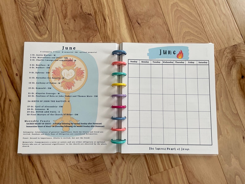 May include: A June calendar page with a colorful design. The left side features a religious image of a monstrance with the letters "THS" in the center. The right side is a blank calendar grid with the days of the week labeled. The bottom of the page says "The Sacred Heart of Jesus".