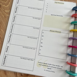 May include: A weekly planner with a white background and black text. The planner includes sections for each day of the week, as well as sections for top priorities, spiritual reminders, and a dinner planner. The planner also includes a quote from St. Anthony of Padua: "Actions speak louder than words. Let your words teach and your actions speak."