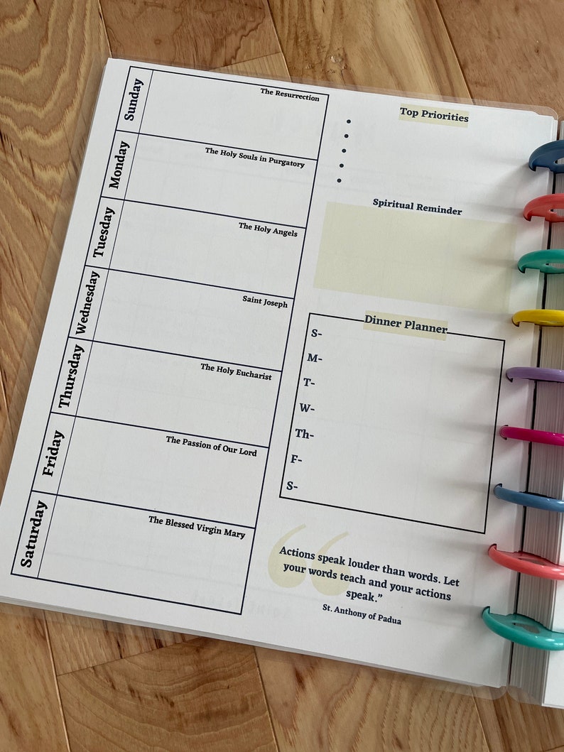 May include: A weekly planner with a white background and black text. The planner includes sections for each day of the week, as well as sections for top priorities, spiritual reminders, and a dinner planner. The planner also includes a quote from St. Anthony of Padua: "Actions speak louder than words. Let your words teach and your actions speak."