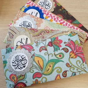 May include: A stack of four colorful envelopes with a floral pattern. Each envelope has a round, silver glitter sticker with Arabic calligraphy that says "Eid Mubarak".