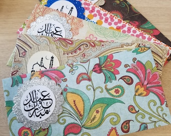 Puede incluir: Una pila de cuatro sobres coloridos con un patrón floral. Cada sobre tiene una pegatina redonda de purpurina plateada con caligrafía árabe que dice "Eid Mubarak".