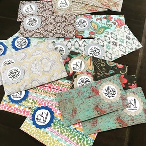 May include: A collection of colorful envelopes with decorative patterns and silver glitter accents. Each envelope features a circular sticker with Arabic calligraphy and the phrase "Eid Mubarak".