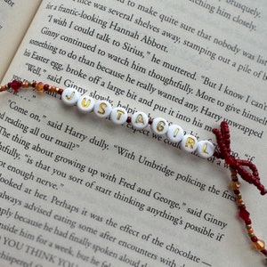 May include: A white and gold beaded bracelet with the word "JUSTAGIRL" spelled out. The bracelet is on a book with red and gold beads.