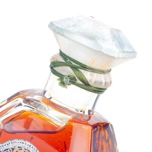 May include: Close-up of a vintage perfume bottle. The bottle has a clear, faceted glass stopper with a green string tied around the neck. The bottle itself is filled with a reddish-brown liquid and has a decorative label.