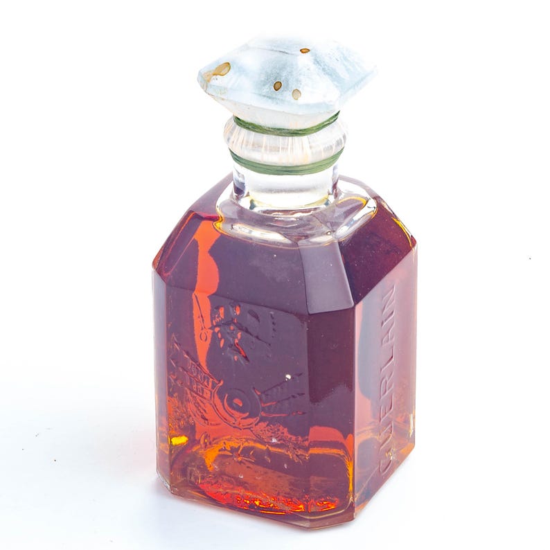 May include: A vintage perfume bottle with a clear, faceted body filled with amber liquid. The bottle has a light blue and white cap and a green ring around the neck. The word "GUERLAIN" is printed on the side of the bottle.