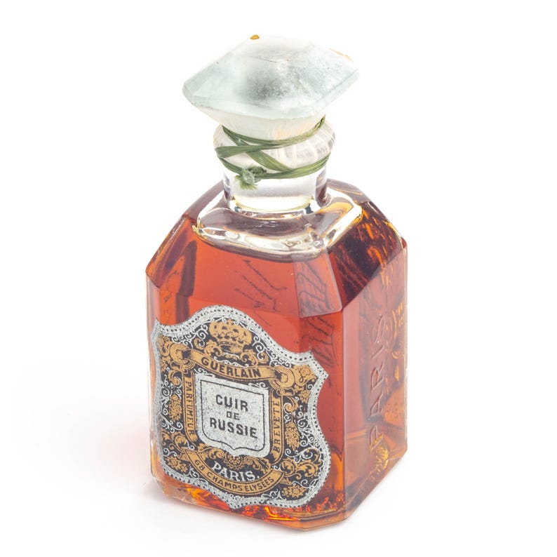 May include: A vintage perfume bottle with a light brown liquid. The bottle has a geometric shape with a decorative label featuring gold and black accents. The bottle is topped with a light green stopper and tied with a green ribbon.