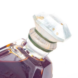 May include: A vintage perfume bottle with a faceted, clear glass stopper. The bottle is filled with a dark amber liquid. The stopper is secured with a green band. The bottle has a geometric shape.