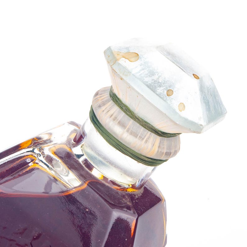 May include: A vintage perfume bottle with a faceted, clear glass stopper. The bottle is filled with a dark amber liquid. The stopper is secured with a green band. The bottle has a geometric shape.