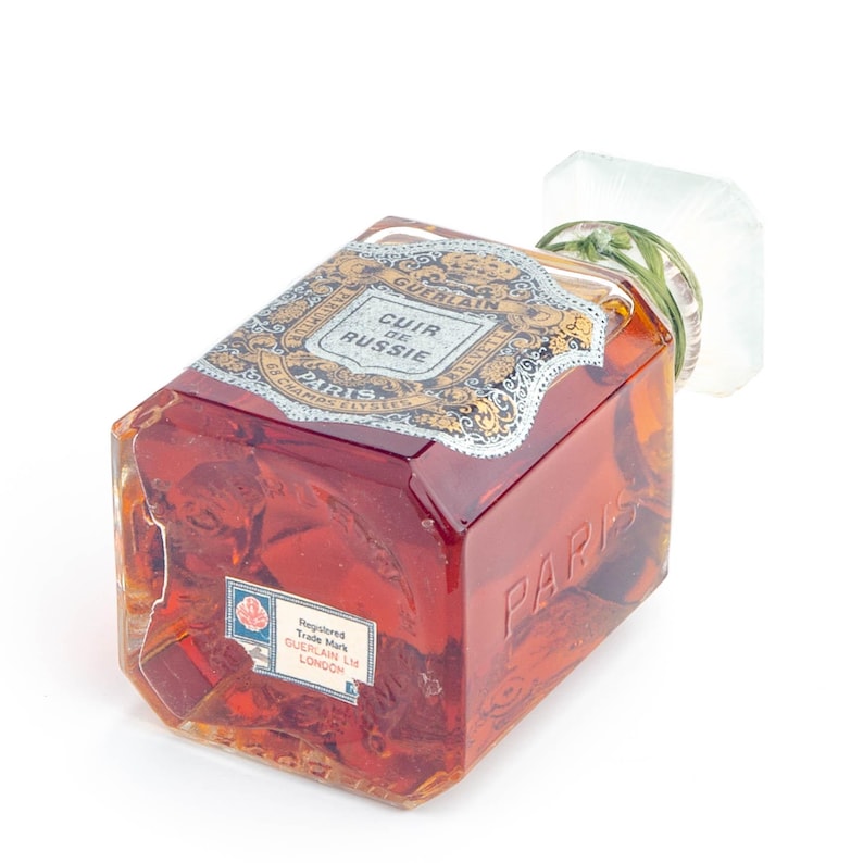 May include: A vintage perfume bottle, rectangular with a clear glass body and a reddish-brown liquid inside. The label reads "Cuir de Russie" and "Paris". The bottle has a white and green stopper.