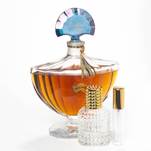 May include: A vintage bottle of Shalimar perfume by Guerlain. The bottle is clear glass with a gold-colored cap and a blue and iridescent stopper. The bottle is sitting on a white surface next to a smaller, clear glass bottle with a gold-colored cap.