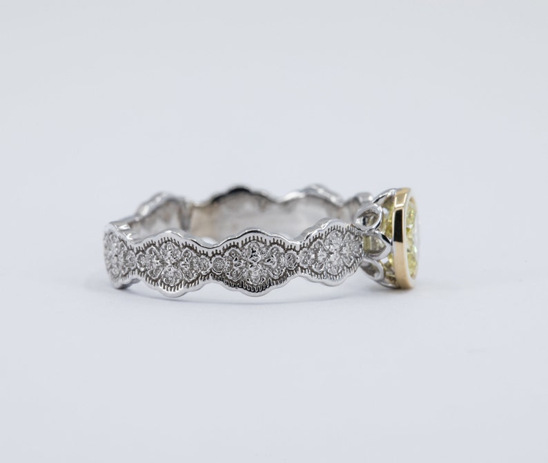 May include: A delicate, white gold engagement ring with a yellow gold band and a round, bezel-set diamond. The band is adorned with a scalloped design and small diamonds.