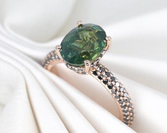 May include: A rose gold ring with a large, oval-cut green gemstone. The band is encrusted with small black stones. The ring is set against a white, textured background, highlighting the jewelry's sparkle and design.