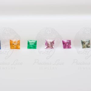 May include: A row of seven square-shaped gemstones in different colors, including blue, orange, green, pink, and light blue. The gemstones are arranged in a line on a white surface. Precious Lace Jewelry is printed below the gemstones.