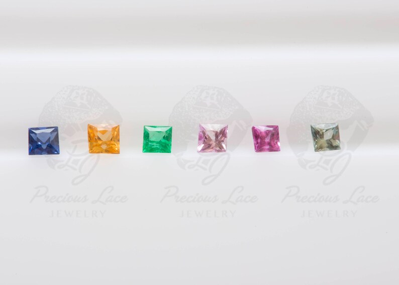 May include: A row of seven square-shaped gemstones in different colors, including blue, orange, green, pink, and light blue. The gemstones are arranged in a line on a white surface. Precious Lace Jewelry is printed below the gemstones.