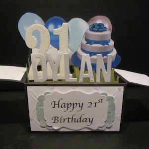 Könnte beinhalten: Eine Pop-up-Geburtstagskarte mit einem dreistöckigen Kuchen, blauen Bändern, Luftballons und dem Namen "Dylan" mit der Zahl "21". Auf der Karte steht "Happy 21st Birthday" auf der Vorderseite.