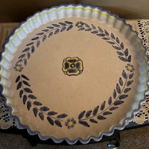 May include: A round, beige ceramic serving dish with a scalloped edge. The dish features a floral and leaf design in blue and yellow. The rim has a blue and yellow trim.