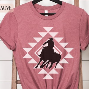 Pu&ograve; includere: Una t-shirt color malva sfumato con la silhouette di un cowboy che cavalca un toro infuriato all'interno di uno schema geometrico.