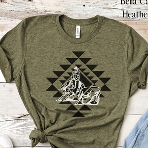 May include: A green Bella Canvas 3001 t-shirt with a white graphic of a cowgirl riding a horse and a steer in a geometric pattern. The text "Heather Olive" is visible in the top right corner of the image.