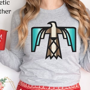 Puede incluir: Una camiseta gris de manga larga con un diseño de águila geométrica turquesa y marrón. La camiseta la lleva puesta una persona que lleva pantalones con estampado rojo y blanco.