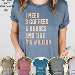 Op de afbeelding: Een blauw Bella Canvas 3001 T-shirt met de tekst "I need 3 coffees 6 horses and like $18 million" erop gedrukt. Het shirt is te zien in vijf verschillende kleuren: heide olijf, heide mauve, donker heide teal, donker heide en atletisch heide.