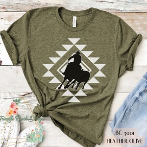 Pu&ograve; includere: Una maglietta verde con un design geometrico bianco e una silhouette nera di un cowboy a cavallo. Il testo "BC 3001 HEATHER OLIVE" &egrave; in basso nell'immagine.