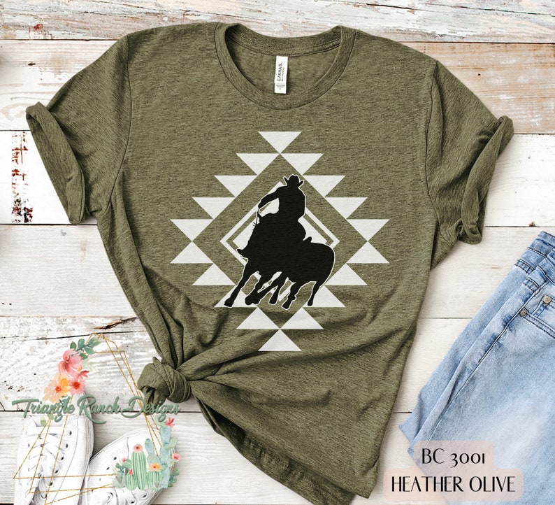 Pu&ograve; includere: Una maglietta verde con un design geometrico bianco e una silhouette nera di un cowboy a cavallo. Il testo "BC 3001 HEATHER OLIVE" &egrave; in basso nell'immagine.