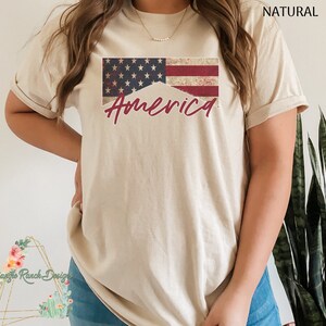 May include: A beige t-shirt with a distressed American flag design and the word "America" in red script.