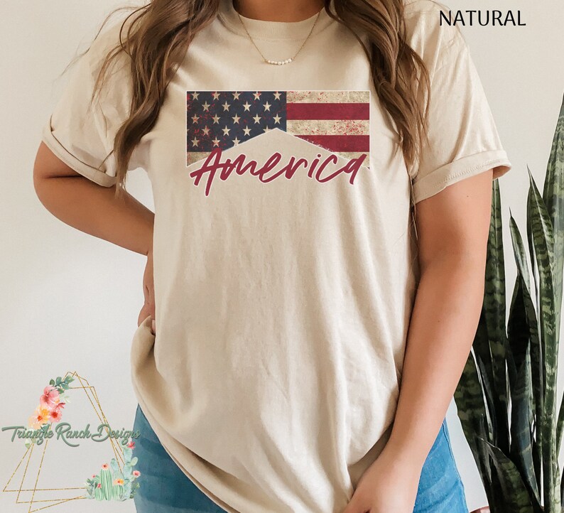 May include: A beige t-shirt with a distressed American flag design and the word "America" in red script.