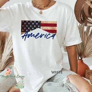 May include: White t-shirt with a distressed American flag design and the word "America" in blue lettering.