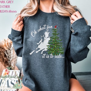 May include: Dark heather grey sweatshirt with a white silhouette of a person riding a bucking horse in front of a Christmas tree with a Santa hat on top. The text reads "Oh what fun it is to ride..."
