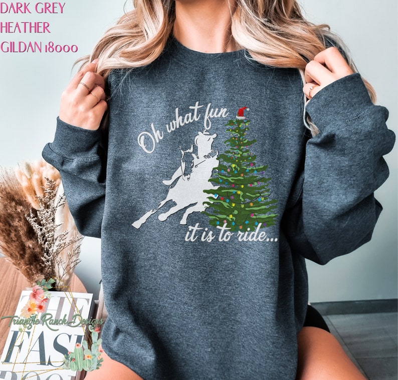 May include: Dark heather grey sweatshirt with a white silhouette of a person riding a bucking horse in front of a Christmas tree with a Santa hat on top. The text reads "Oh what fun it is to ride..."