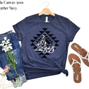May include: A navy blue Bella Canvas 3001 t-shirt with a white graphic of a cowgirl riding a horse and a steer in a geometric pattern. The shirt is on a white background with a pair of brown sandals.