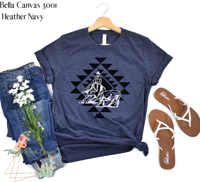 May include: A navy blue Bella Canvas 3001 t-shirt with a white graphic of a cowgirl riding a horse and a steer in a geometric pattern. The shirt is on a white background with a pair of brown sandals.