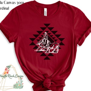 May include: Red Bella Canvas 3001 t-shirt with a white silhouette of a cowboy riding a horse and herding cattle in a geometric pattern.