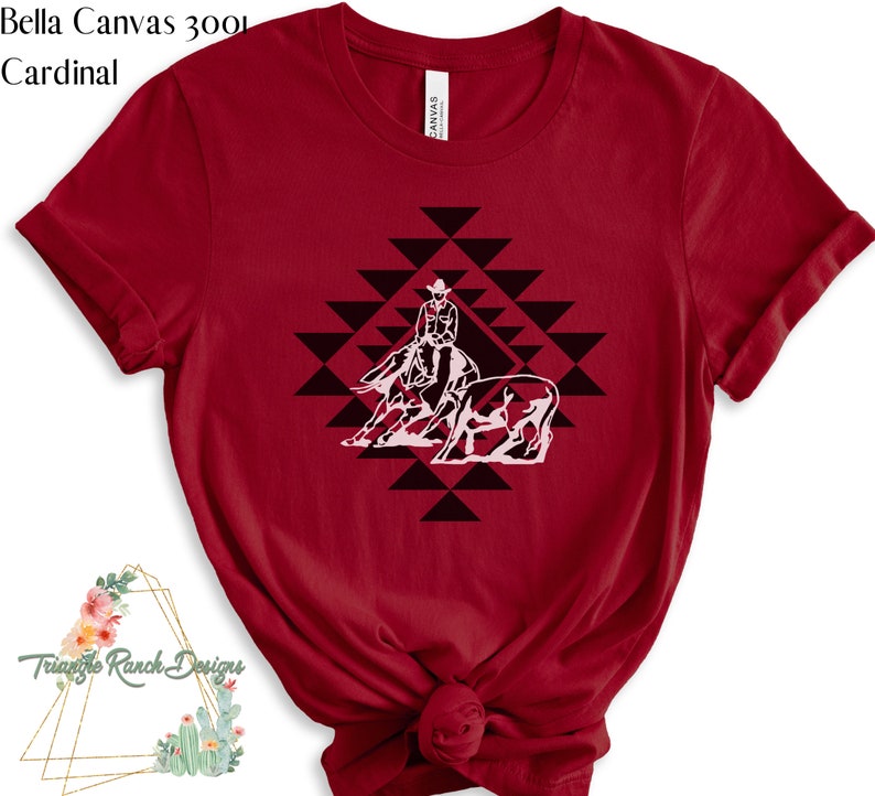 May include: Red Bella Canvas 3001 t-shirt with a white silhouette of a cowboy riding a horse and herding cattle in a geometric pattern.