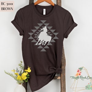 Pu&ograve; includere: Una maglietta marrone con una silhouette bianca di un cowboy a cavallo. Il design &egrave; centrato sulla maglietta e presenta un motivo geometrico di triangoli grigi.