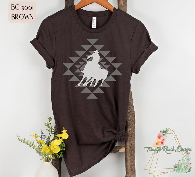 Pu&ograve; includere: Una maglietta marrone con una silhouette bianca di un cowboy a cavallo. Il design &egrave; centrato sulla maglietta e presenta un motivo geometrico di triangoli grigi.