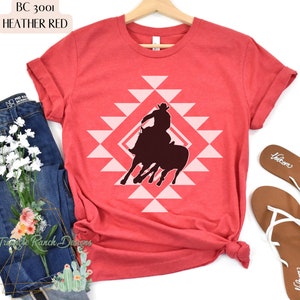 Pu&ograve; includere: Una t-shirt color rosso heather con una silhouette di un cowboy che cavalca un toro selvaggio. Il design &egrave; centrato sulla t-shirt e presenta un motivo geometrico rosa.