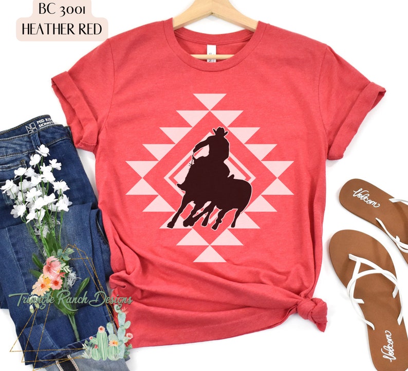 Pu&ograve; includere: Una t-shirt color rosso heather con una silhouette di un cowboy che cavalca un toro selvaggio. Il design &egrave; centrato sulla t-shirt e presenta un motivo geometrico rosa.