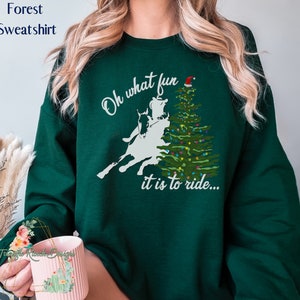 May include: Green forest sweatshirt with a white silhouette of a person riding a horse in front of a Christmas tree with a Santa hat on top. The text reads "Oh what fun it is to ride..."