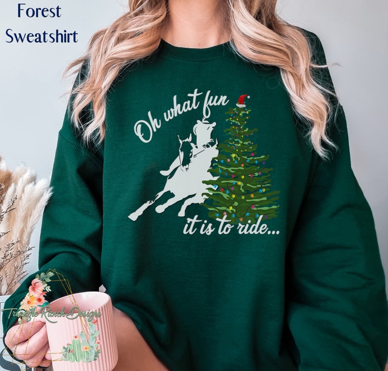 May include: Green forest sweatshirt with a white silhouette of a person riding a horse in front of a Christmas tree with a Santa hat on top. The text reads "Oh what fun it is to ride..."