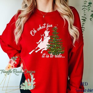 May include: Red sweatshirt with a white graphic of a person riding a horse in front of a Christmas tree. The text on the sweatshirt reads "Oh what fun it is to ride..."