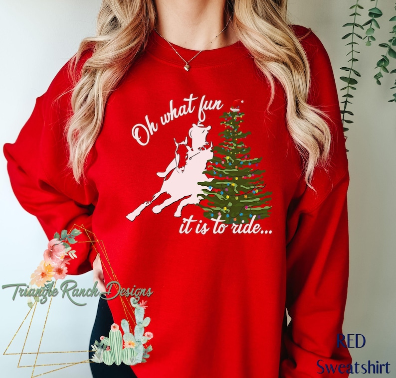 May include: Red sweatshirt with a white graphic of a person riding a horse in front of a Christmas tree. The text on the sweatshirt reads "Oh what fun it is to ride..."