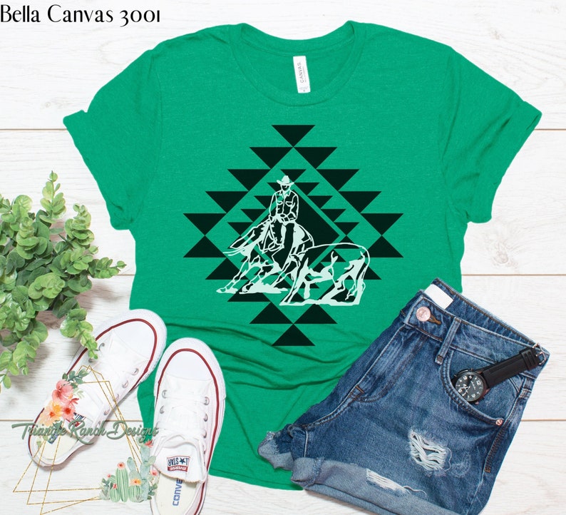 May include: A green Bella Canvas 3001 t-shirt with a white silhouette design of a cowboy riding a horse and a cow. The design is in a geometric pattern of black and white triangles.