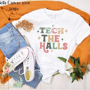 May include: White t-shirt with the text "Tech the Halls" in a retro font with colorful accents and small star designs. The shirt is styled with blue jeans, white sneakers, and a brown cardigan.